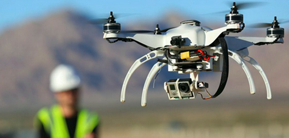 A white drone equipped with a camera flying outdoors against a blurred mountainous background.