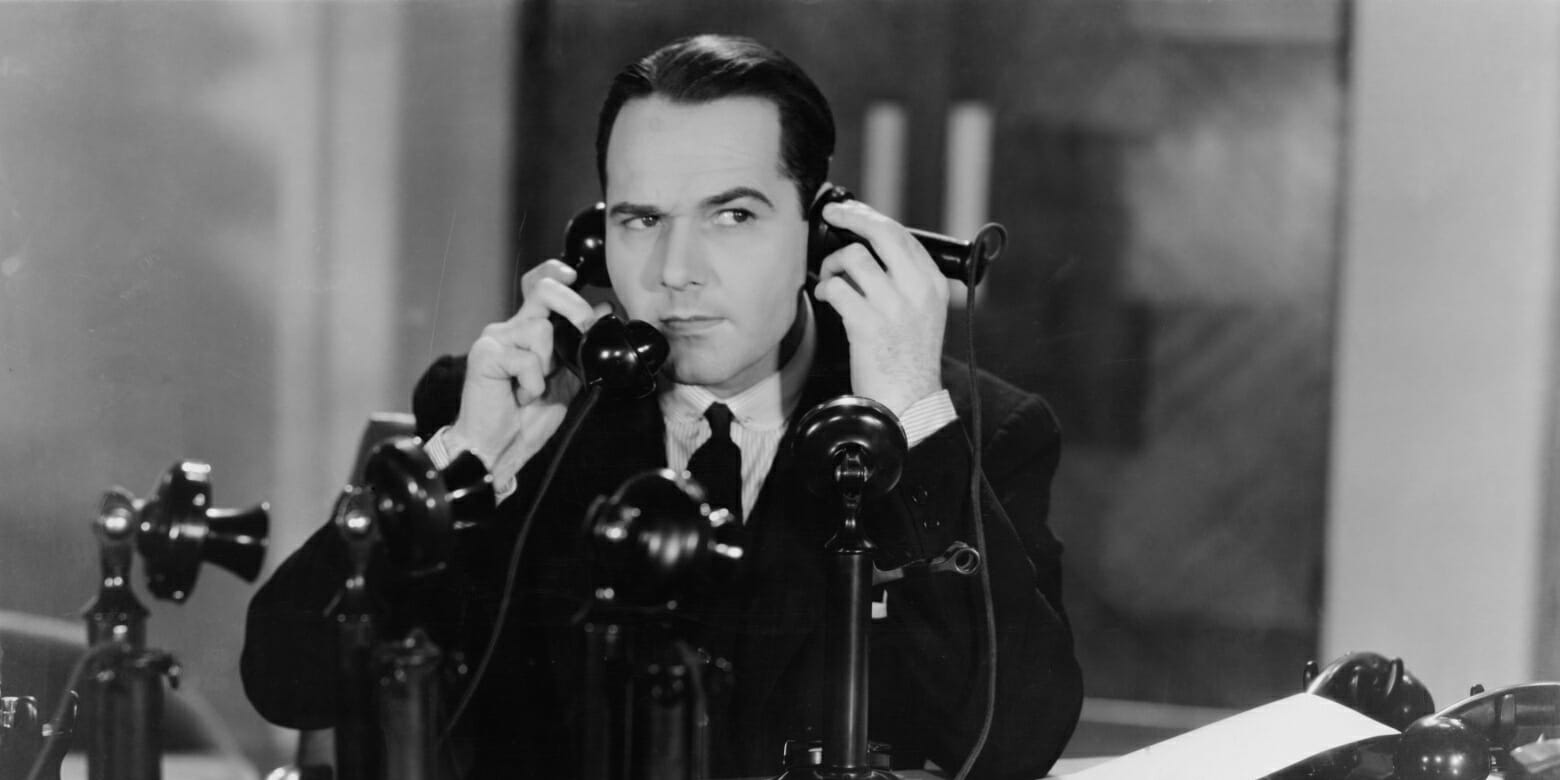 Black and white photo of a man speaking on multiple telephones.