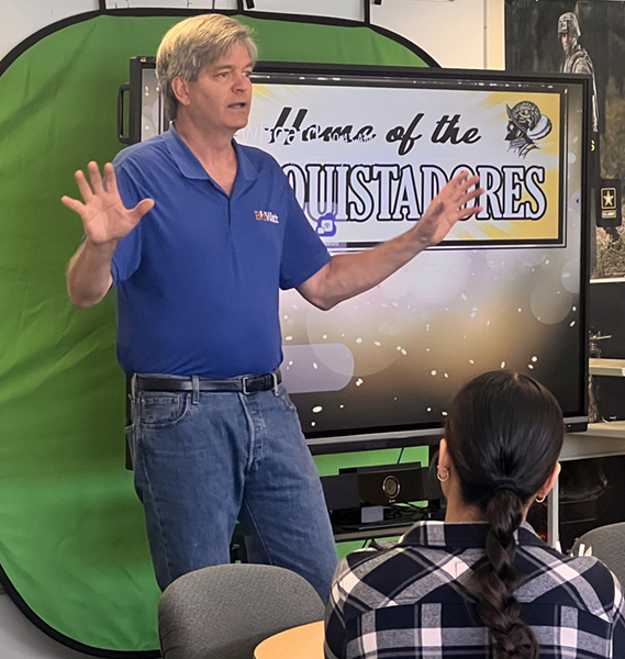 Man giving a presentation in a classroom with a 'Home of the Mustangs' sign.